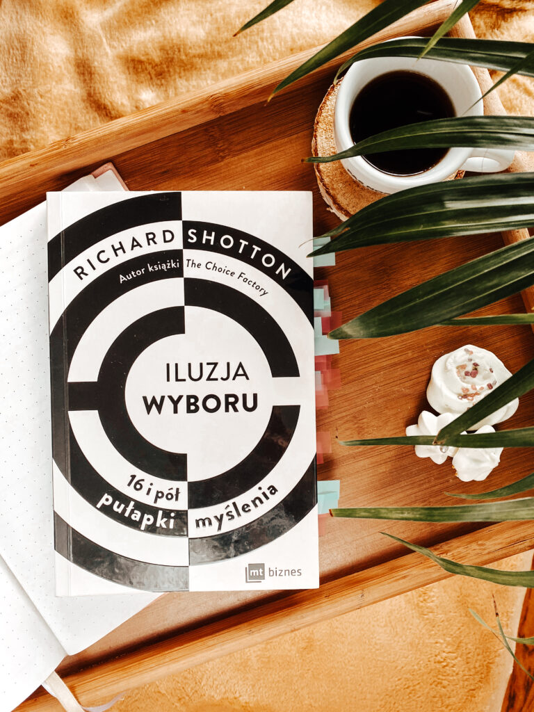 Książka Richarda Shottona "Iluzja wyboru" na podkładce drewnianej, obok kawy i ciastek bezowych.